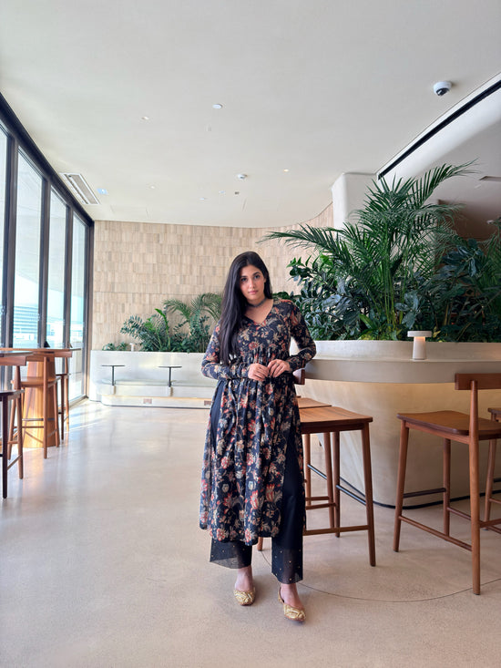 A woman stands in a modern indoor cafe or lounge setting wearing a long-sleeved floral print tunic dress with matching wide-leg pants. The dress features a dark navy base with pink, orange, and green floral patterns.